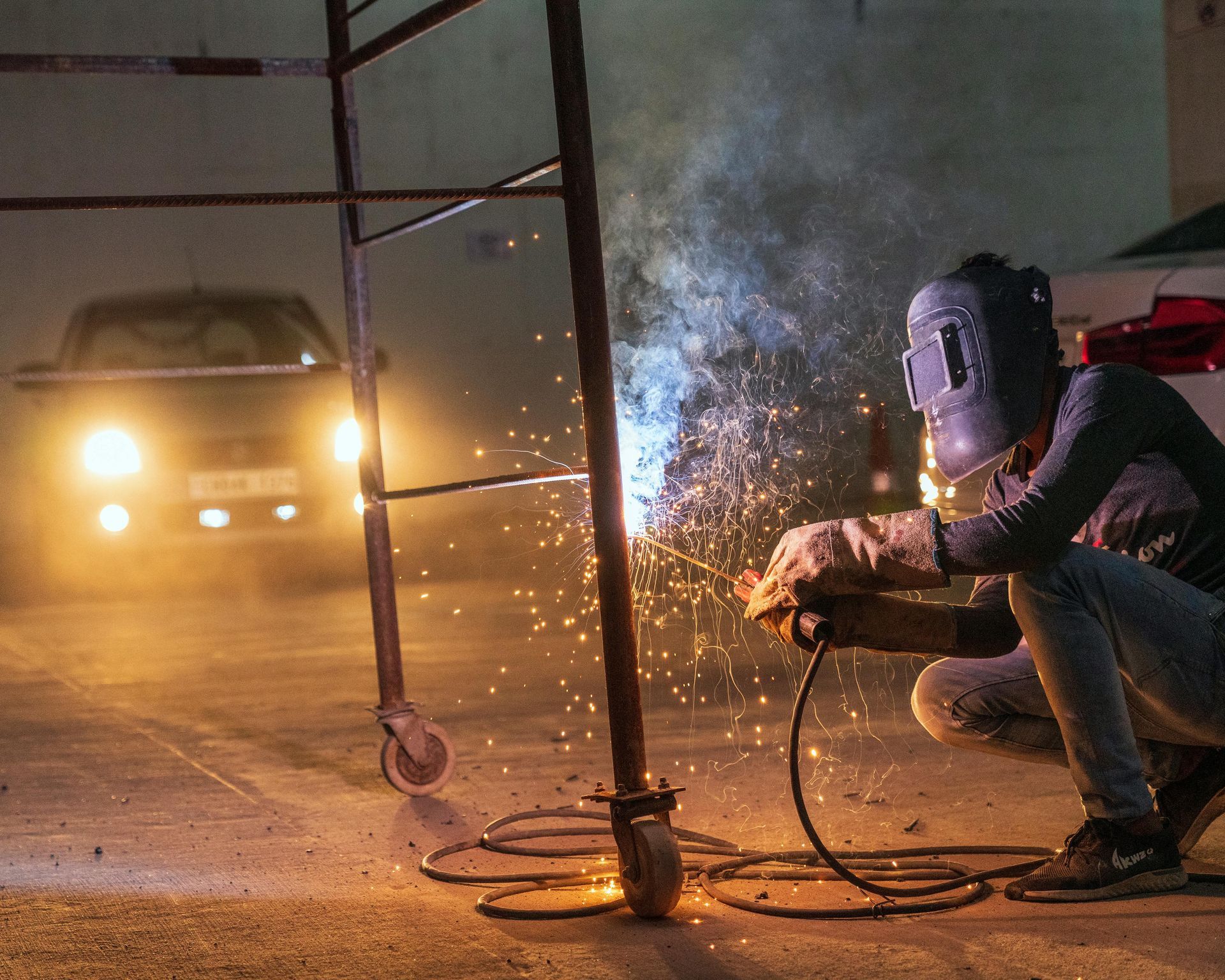 Welder in protective mask using a torch on metal scaffolding; sparks fly in the dark.