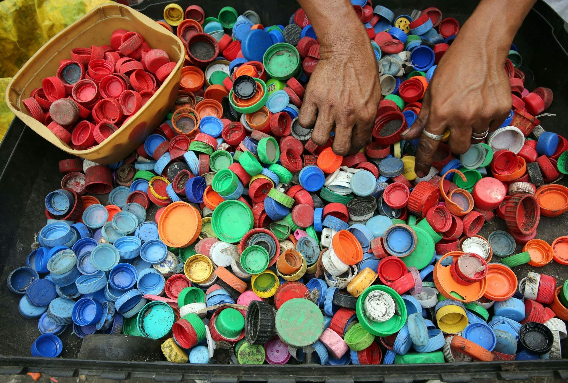 Hands sorting colorful plastic bottle caps in a container; small basket of red caps on left.