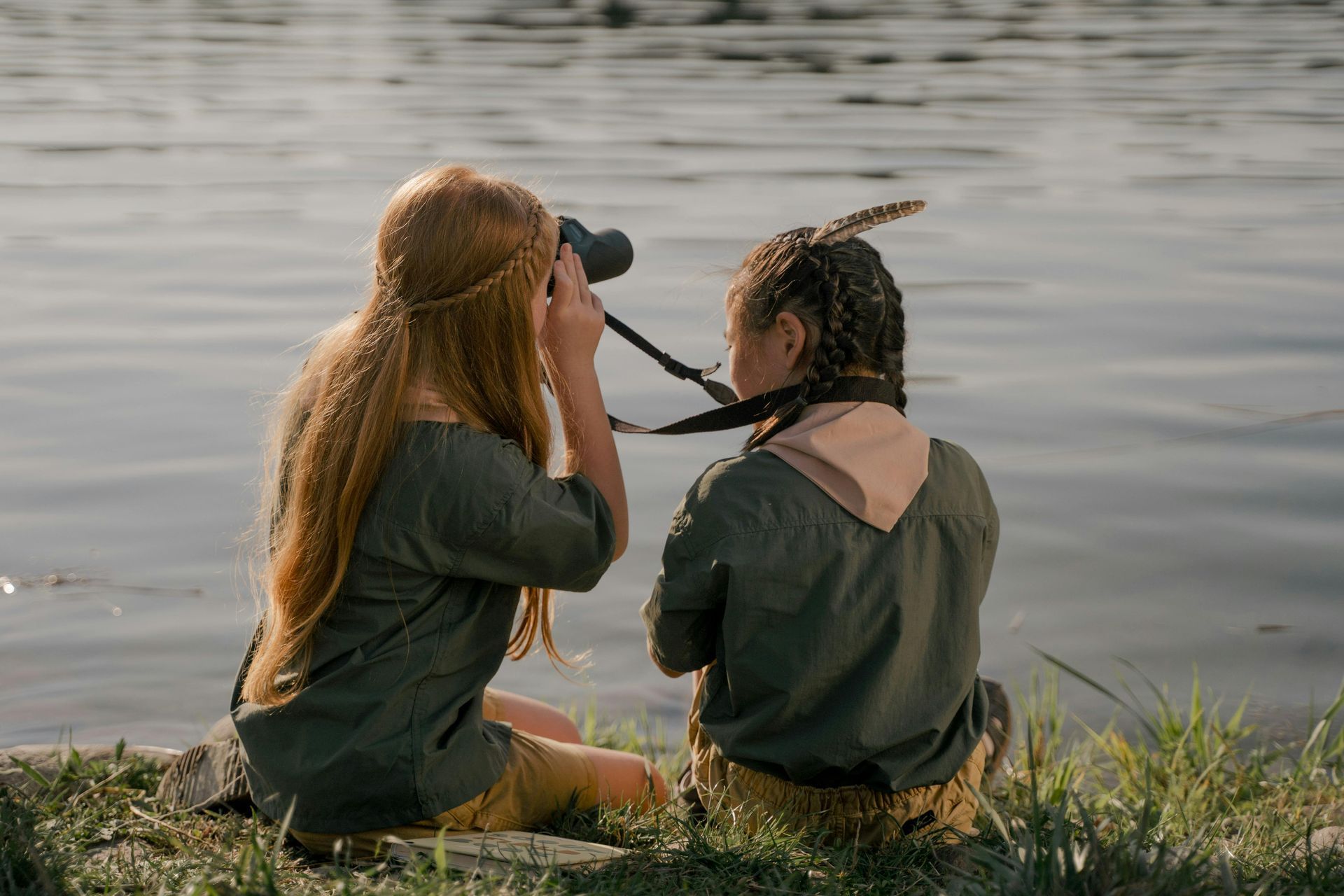 Two people by water, one using binoculars, the other with a feather in their hair, both facing the water.