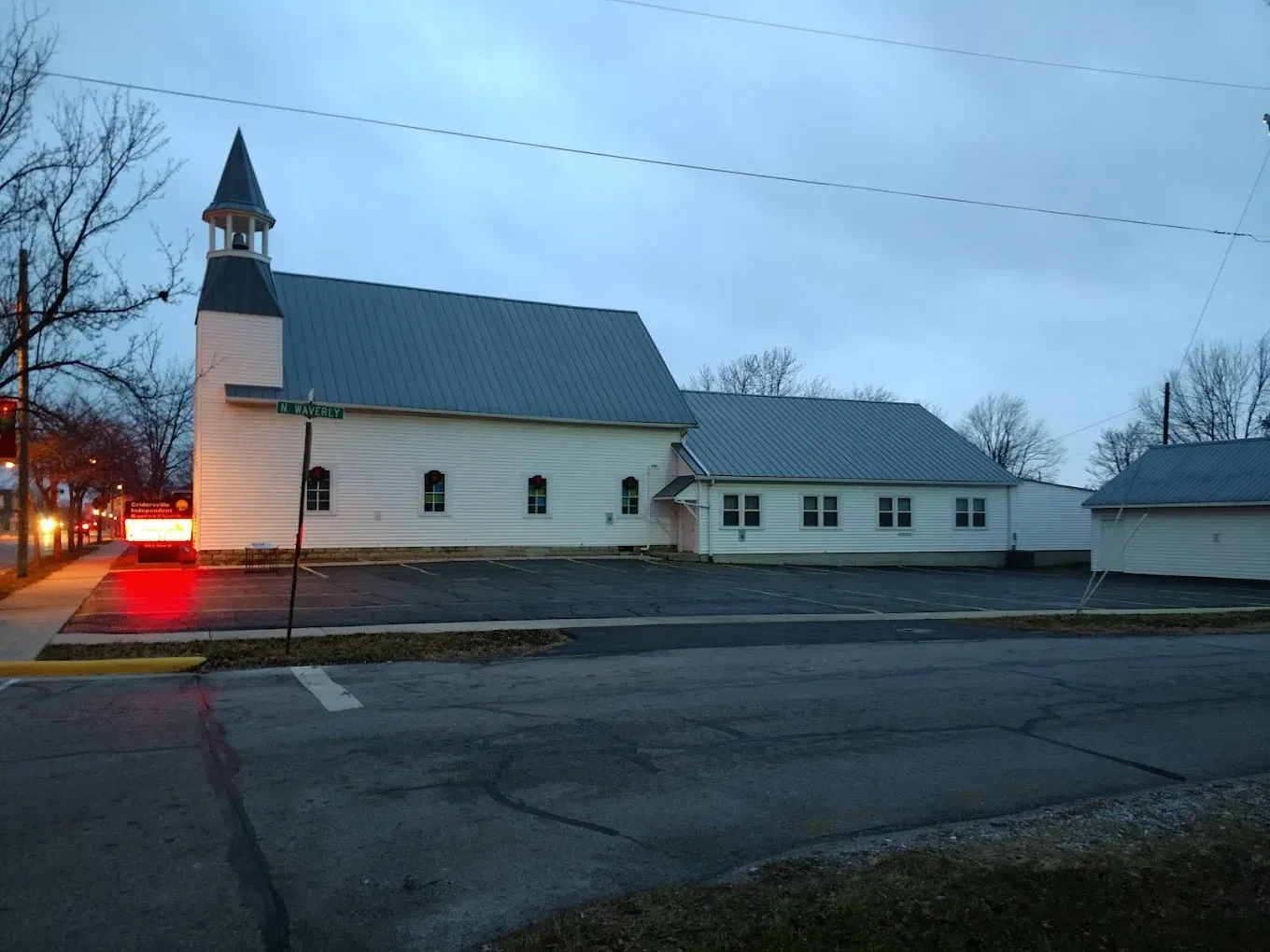 White church building with steeple, gray metal roof, and street in front, dusk setting.