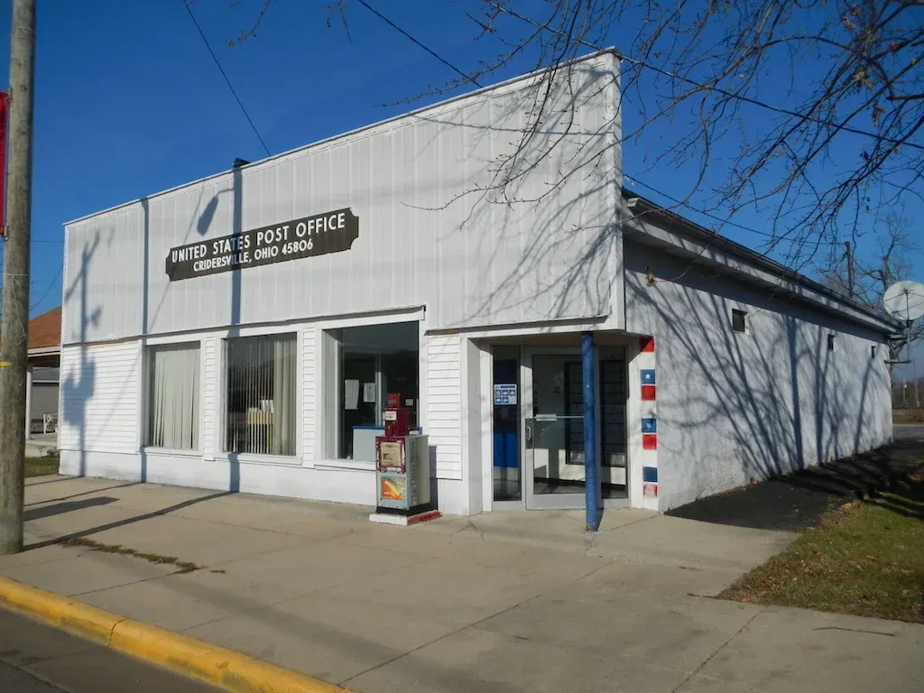 White post office building with blue and red details, sidewalk in front, sunny day.