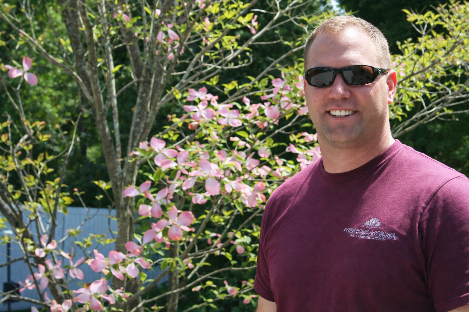 Man in sunglasses and maroon shirt standing outdoors beside pink blossoms and green foliage