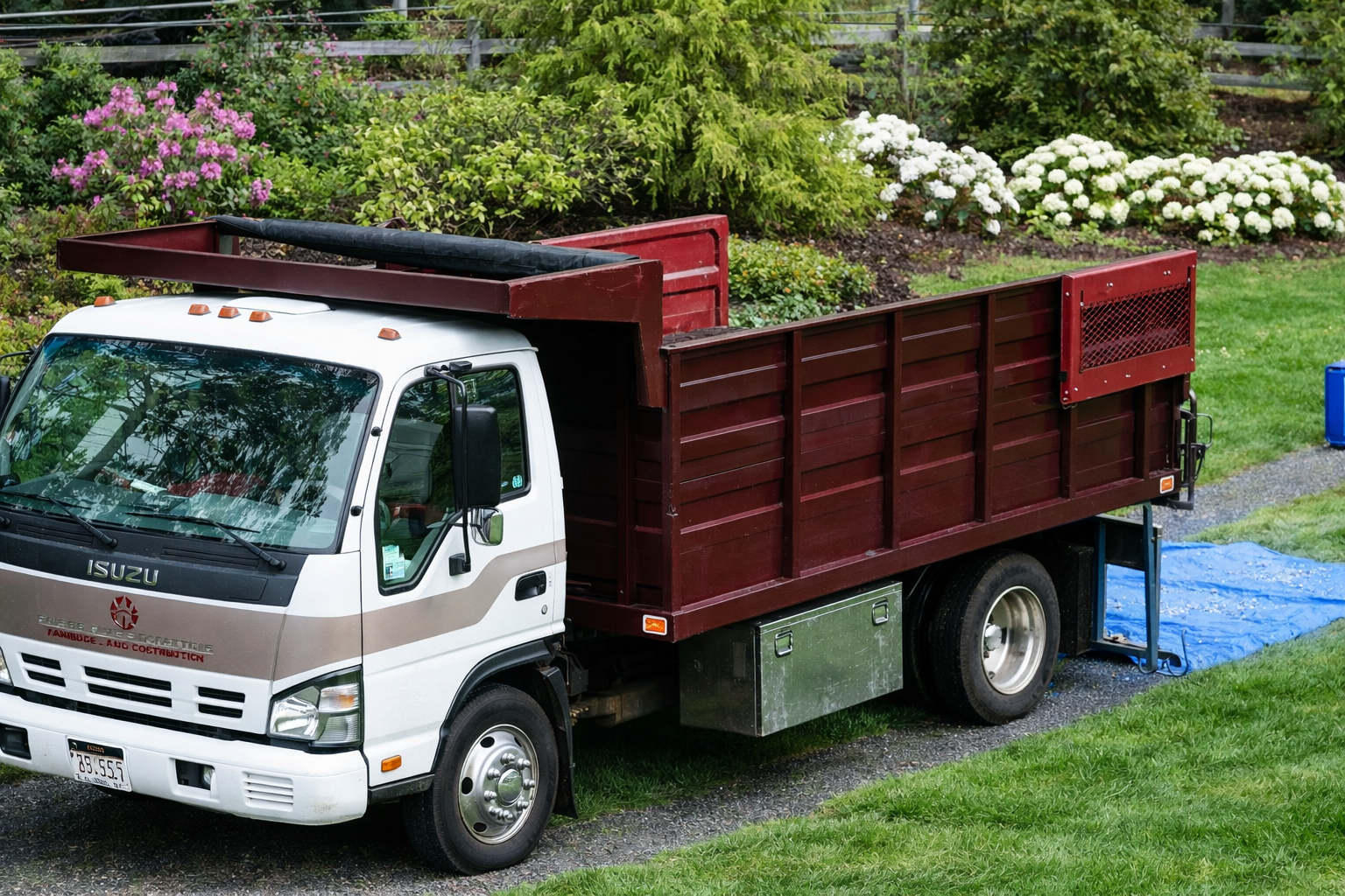 White flatbed truck loaded with red wooden panels, parked on a grassy slope with flowers behind it