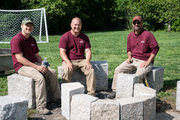 Three people in maroon shirts sit on stone blocks in a grassy park near a soccer goal.