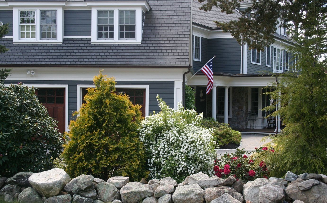 Gray house with white trim, front porch, American flag, and landscaped rocks and shrubs.