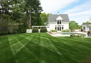 Lush green striped lawn leading to a white house with a pool and surrounding trees