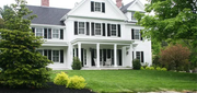 White two-story house with black shutters, front porch, and green lawn under trees