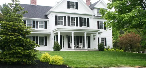 White two-story house with black shutters, front porch, and green lawn under trees