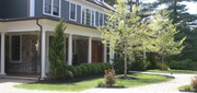 Front porch of a brick house with white columns, shrubs, and a sunlit driveway