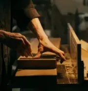Hands shaping wood on a bench under a desk lamp in a workshop