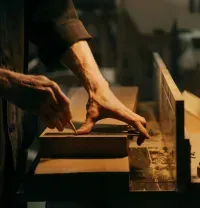 Hands shaping wood on a bench under a desk lamp in a workshop
