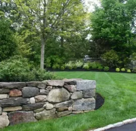 Stone retaining wall beside a manicured green lawn and trees in a landscaped yard