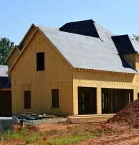 Partially built house with yellow walls and gray roof, surrounded by construction materials and dirt piles