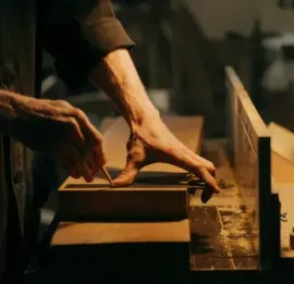 Hands working with wood at a bench under a lamp in a dim workshop