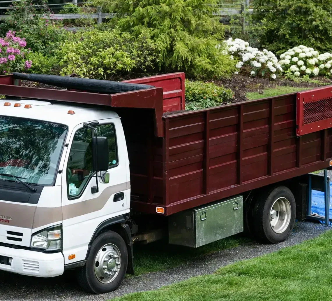 White flatbed truck loaded with red wooden panels, parked on a grassy slope with flowers behind it