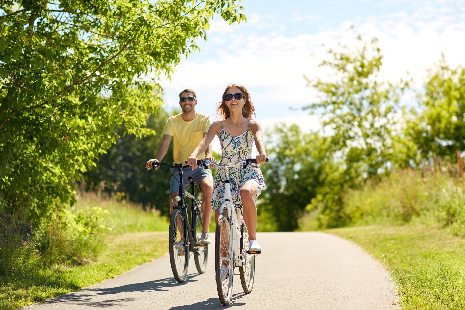 A person in a yellow shirt and another in a patterned dress cycling together on a sunny paved path surrounded by trees.