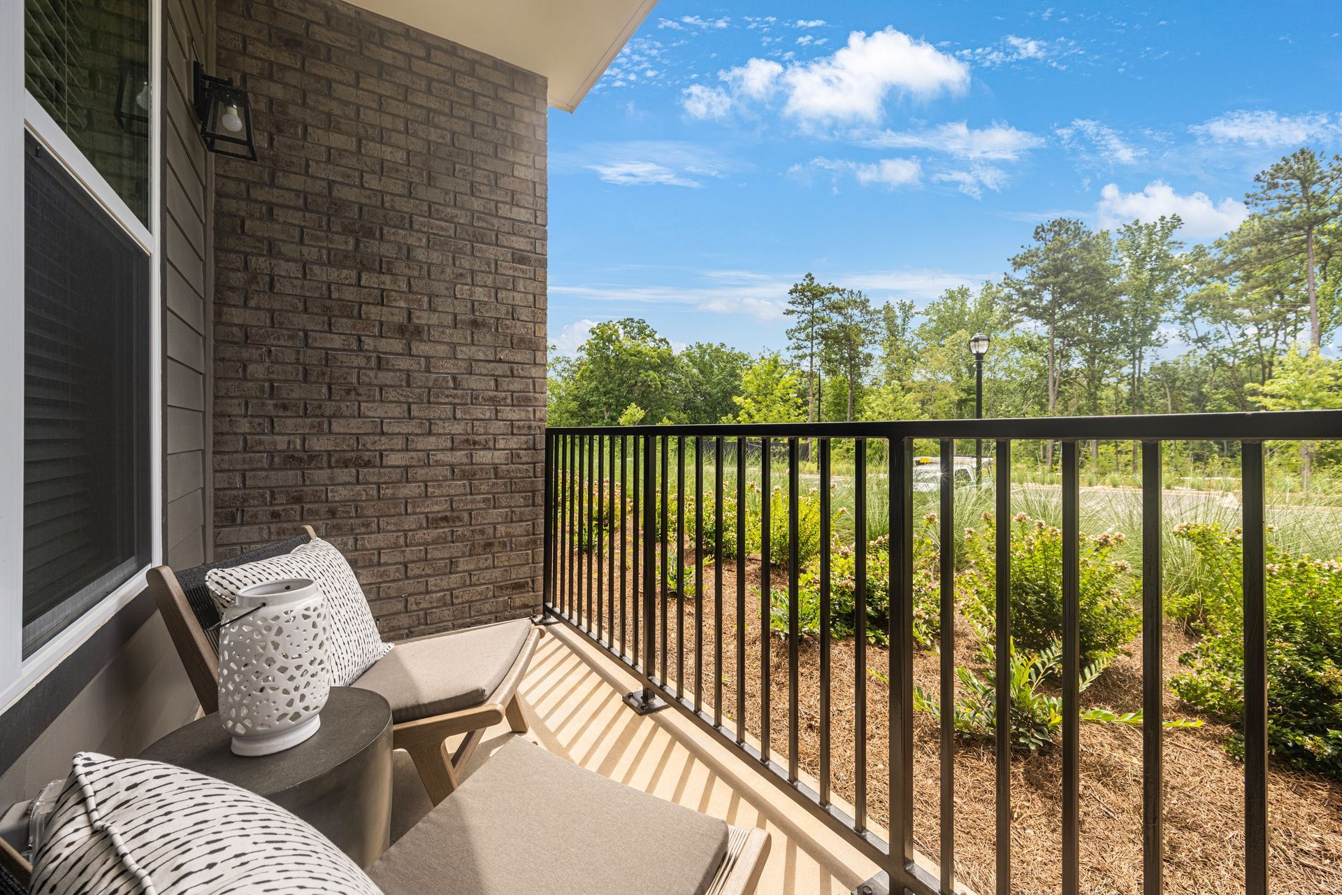 Balcony with brick wall, chairs, and black railing overlooking green trees and a blue sky.