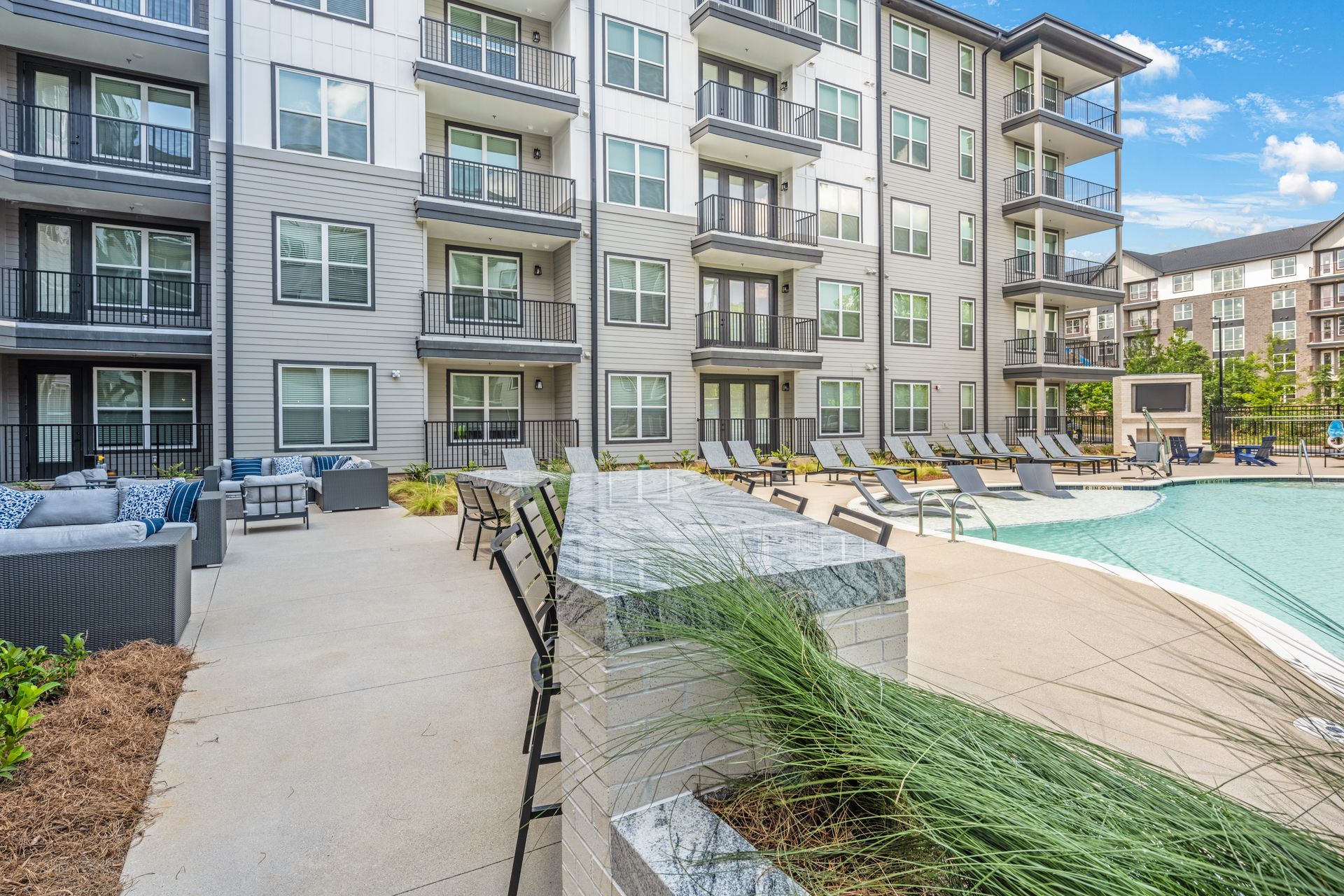 Apartment complex exterior with a pool, patio seating, and balconies under a blue sky.