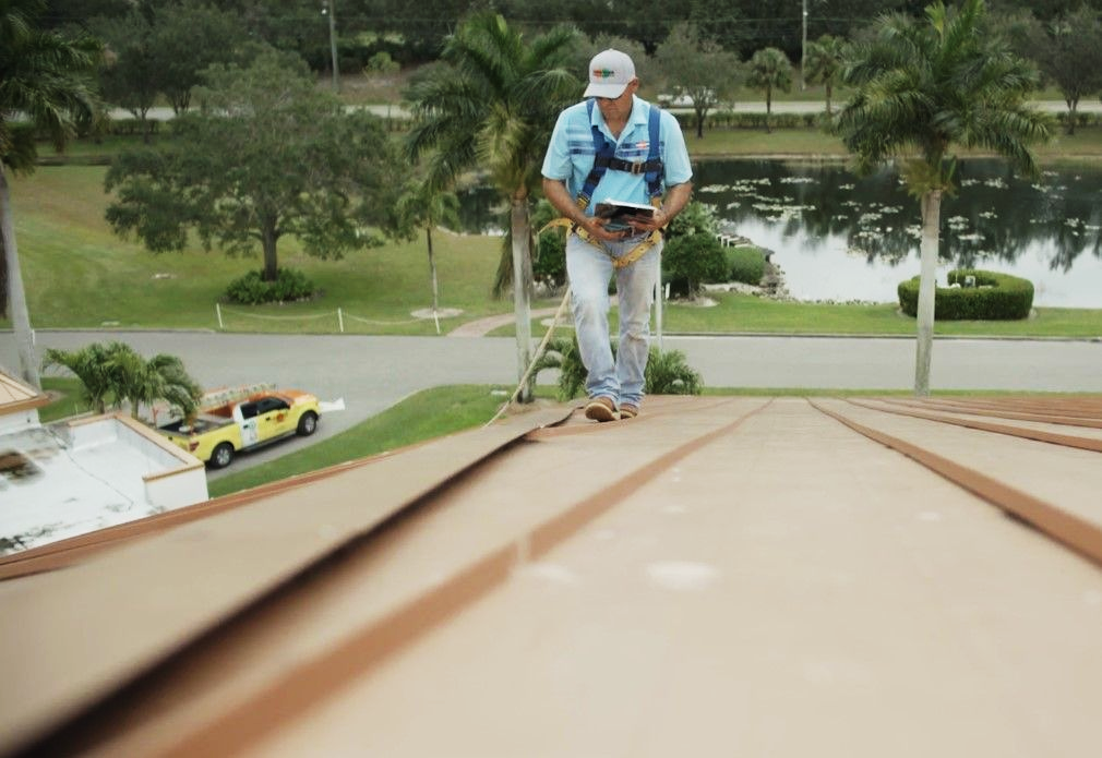 Worker on a rooftop using a rope harness, with palm trees and a pond in the background.