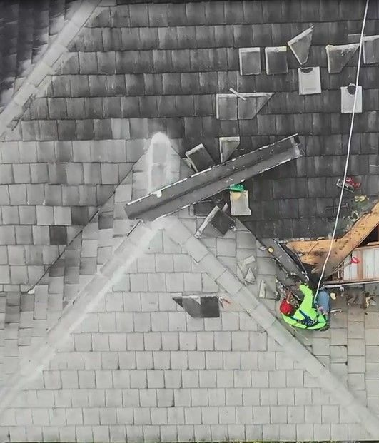 Roofers working on a gray shingled roof beside a skylight and chimney, with safety gear and tools.