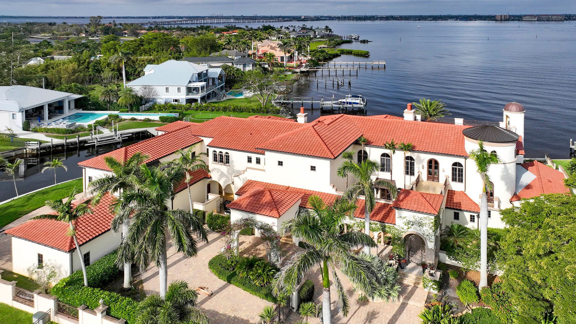 Aerial view of a white villa with red tile roofs beside a waterfront and palm trees