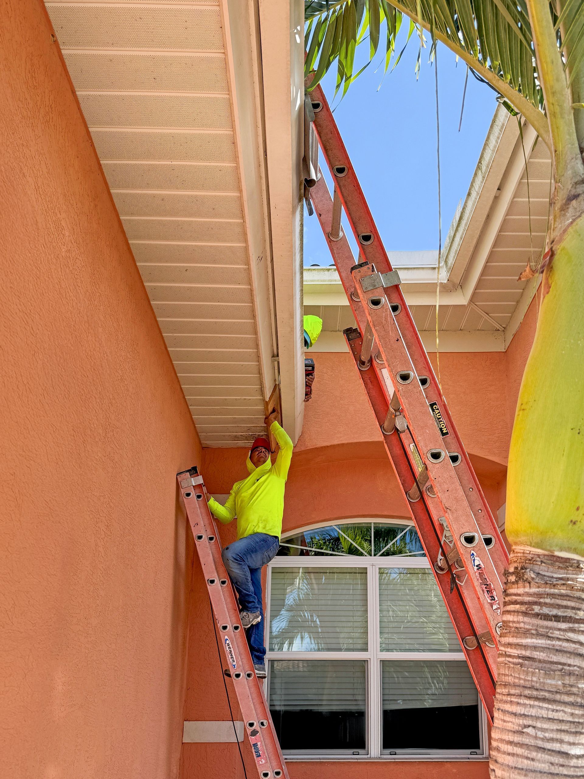 Worker on ladder repairing a peach-colored building’s ceiling near a window and palm fronds