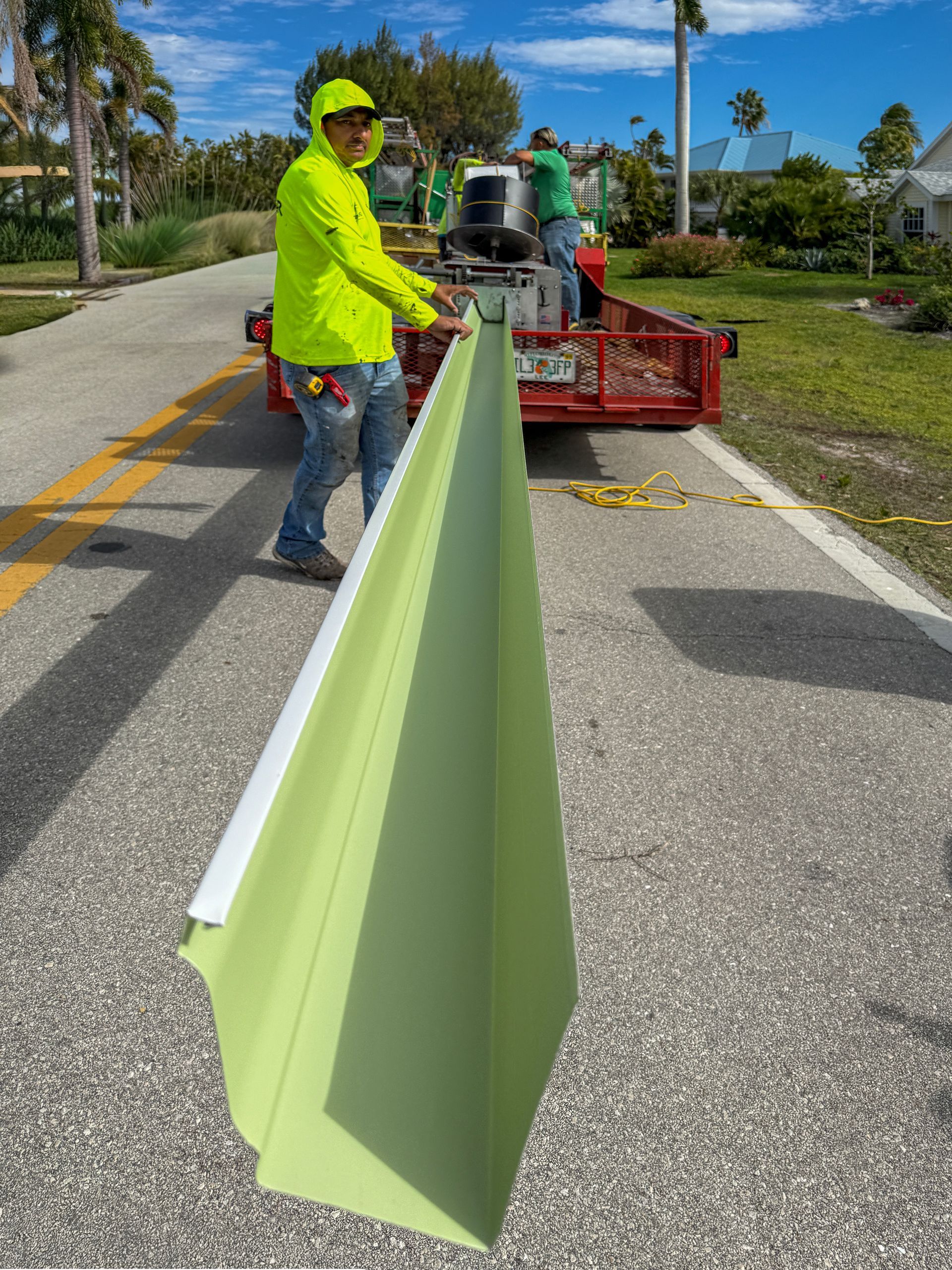 Worker in neon jacket holding a long green board beside a roadside truck under a blue sky