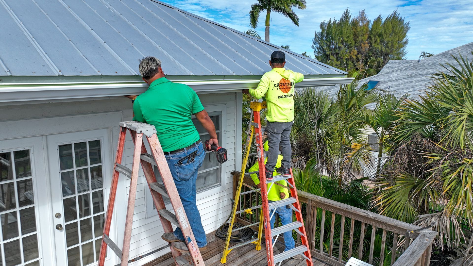 Two workers on ladders cleaning a roof beside a white house on a sunny day