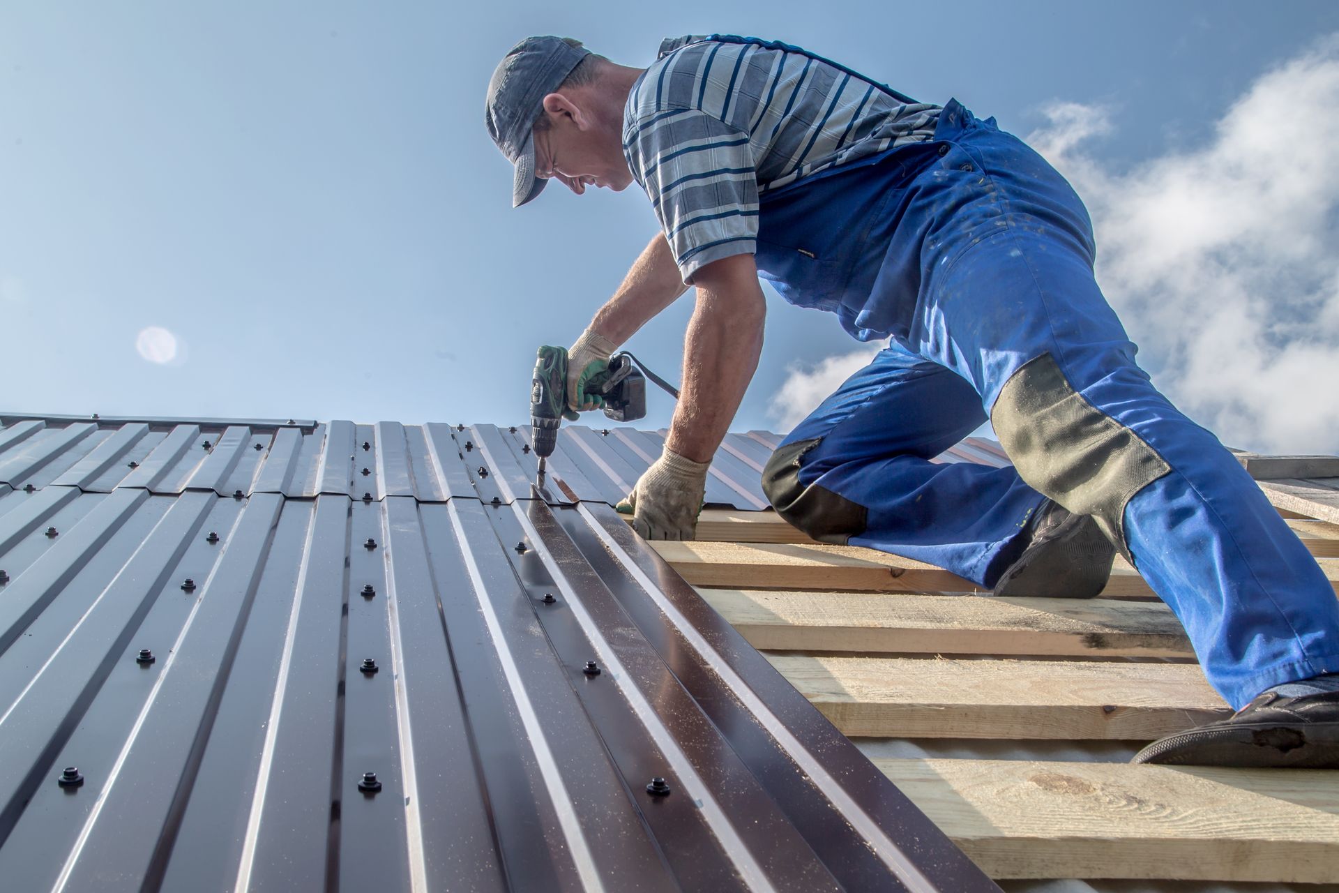Worker installing metal roofing on a house roof, kneeling with a nail gun under a blue sky