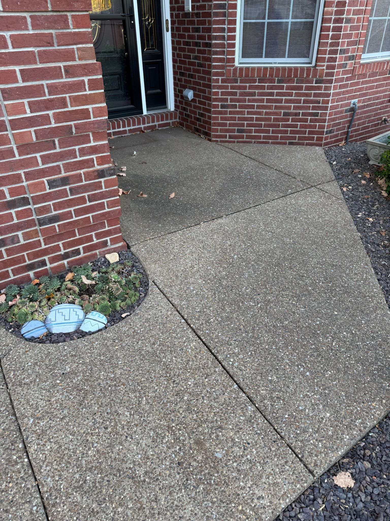 Concrete sidewalk leading to a brick building's entrance. Some landscaping visible on the left side.