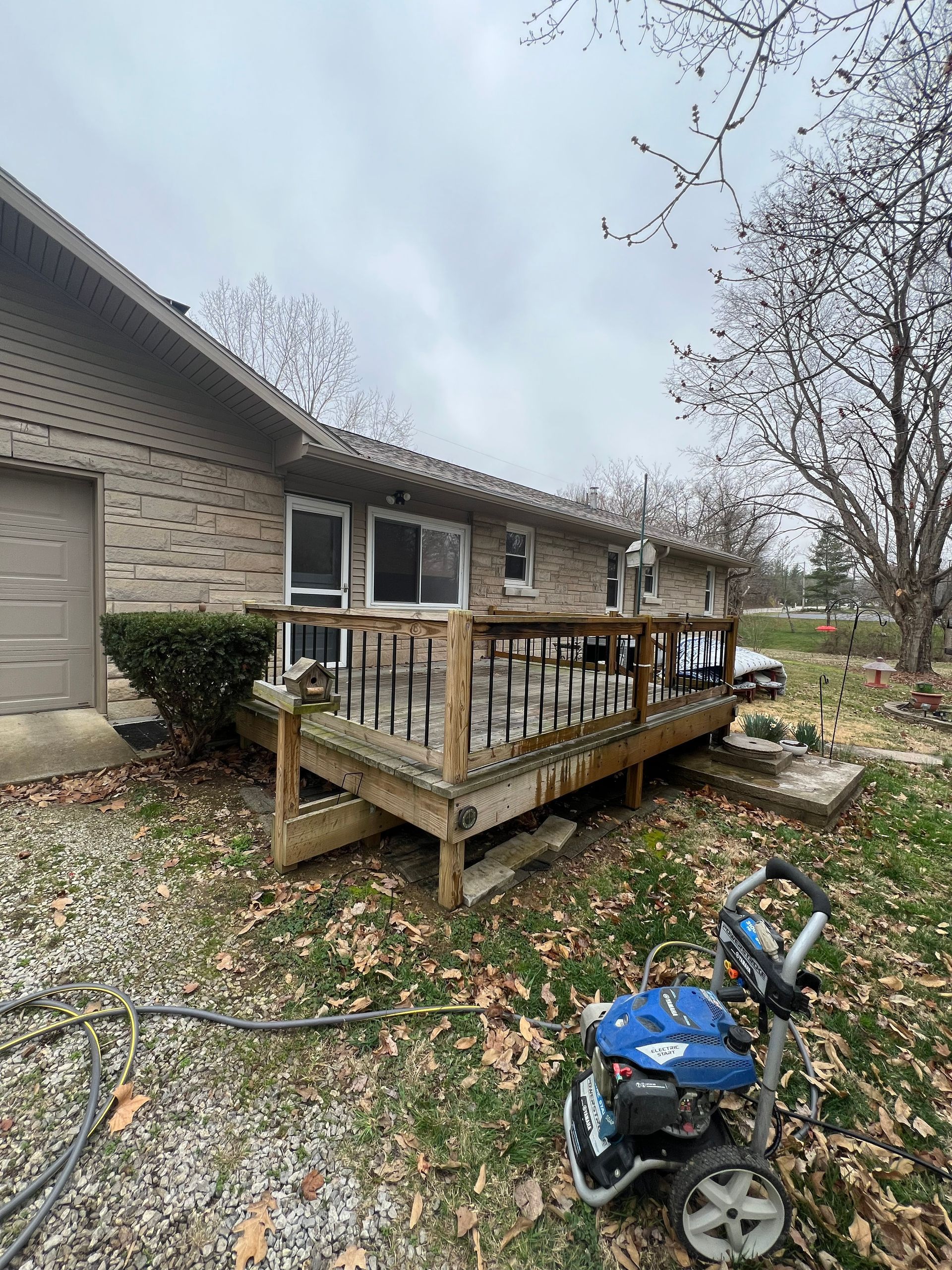 A person pressure washing a weathered wooden deck attached to a brick house on a cloudy day.