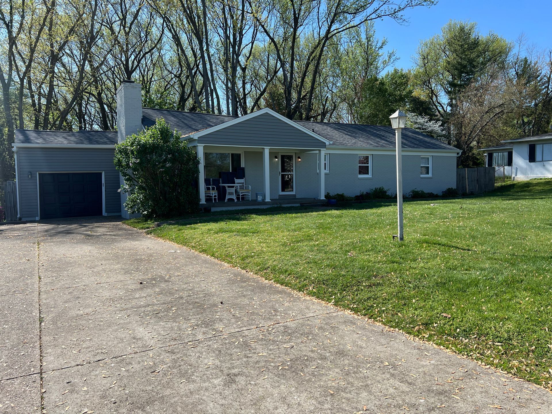 Gray ranch-style house with a driveway, garage, porch, and grassy yard. Trees in the background.
