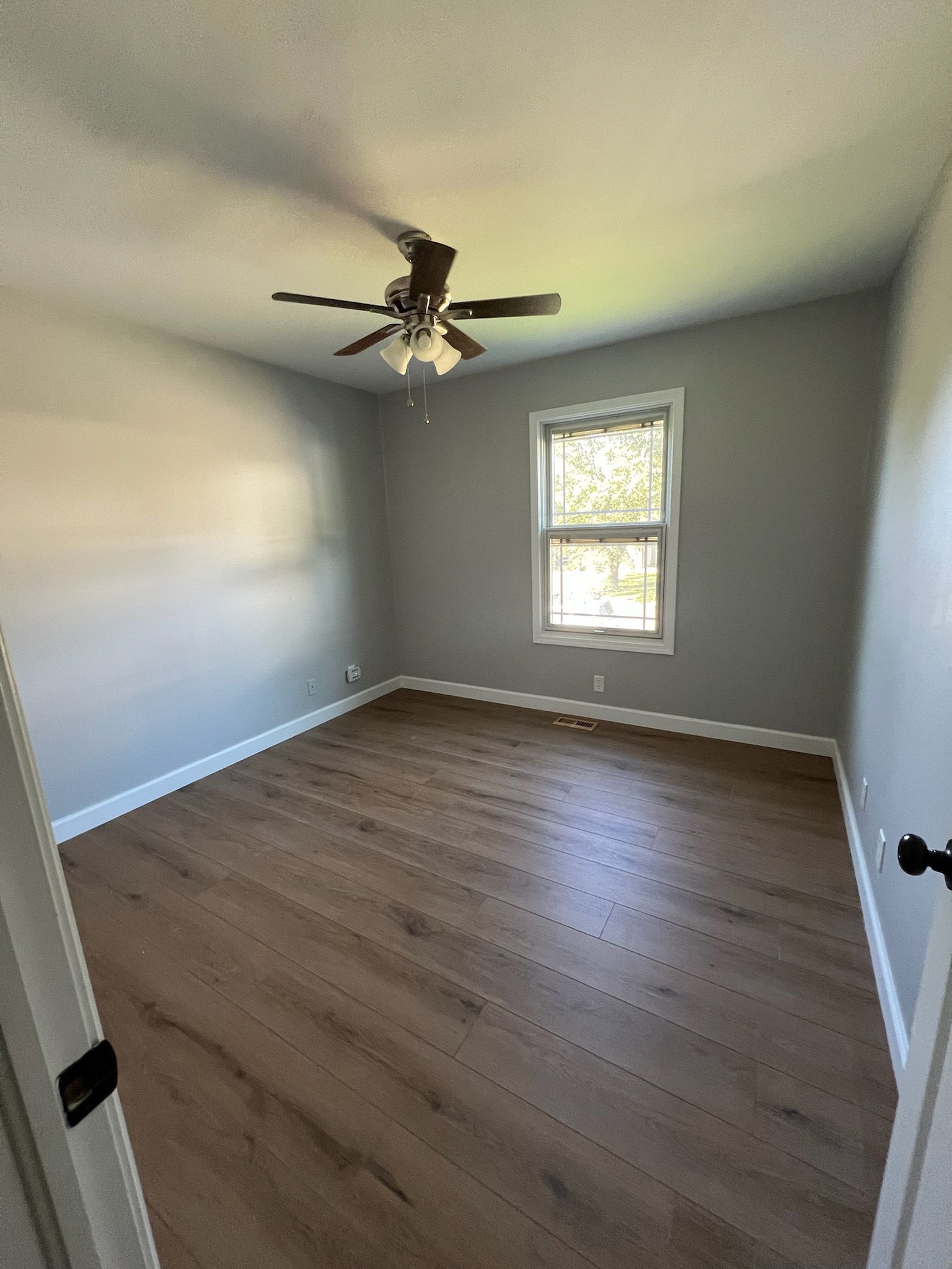 Empty bedroom with gray walls, brown floor, window, and ceiling fan.