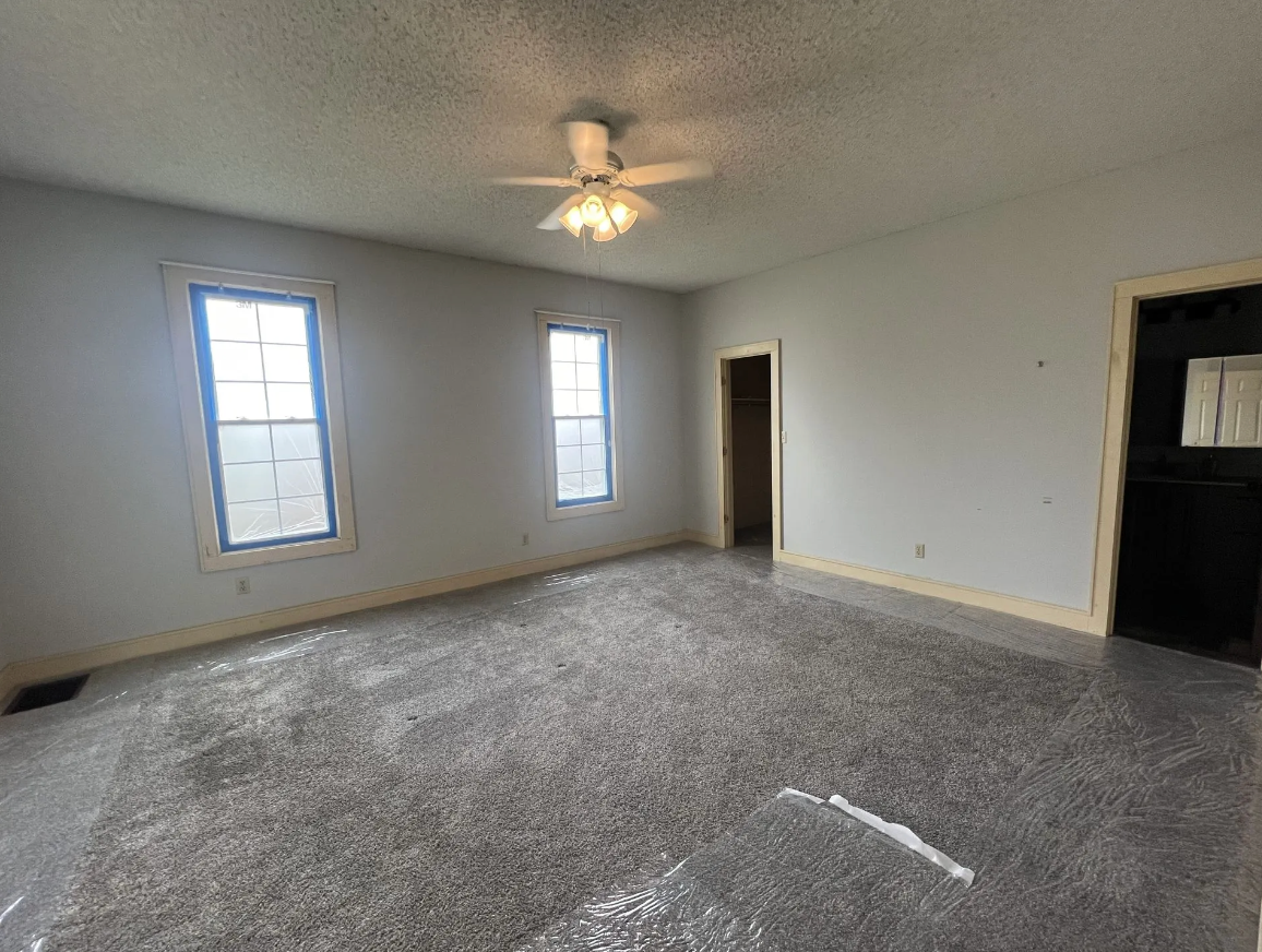 Empty room with gray walls and carpet, two windows, a door, and a ceiling fan.