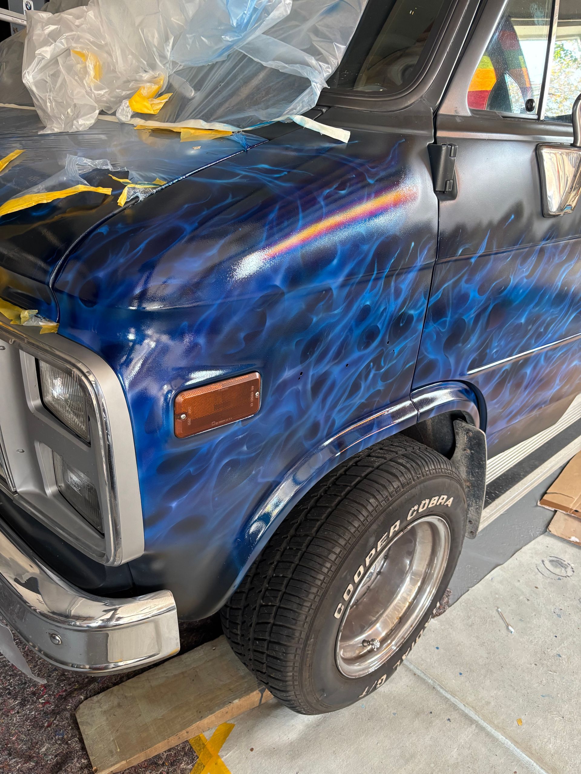 Blauwe pick-up truck met vlammenmotief, geparkeerd in een garage; de ​​linker voorkant is zichtbaar.