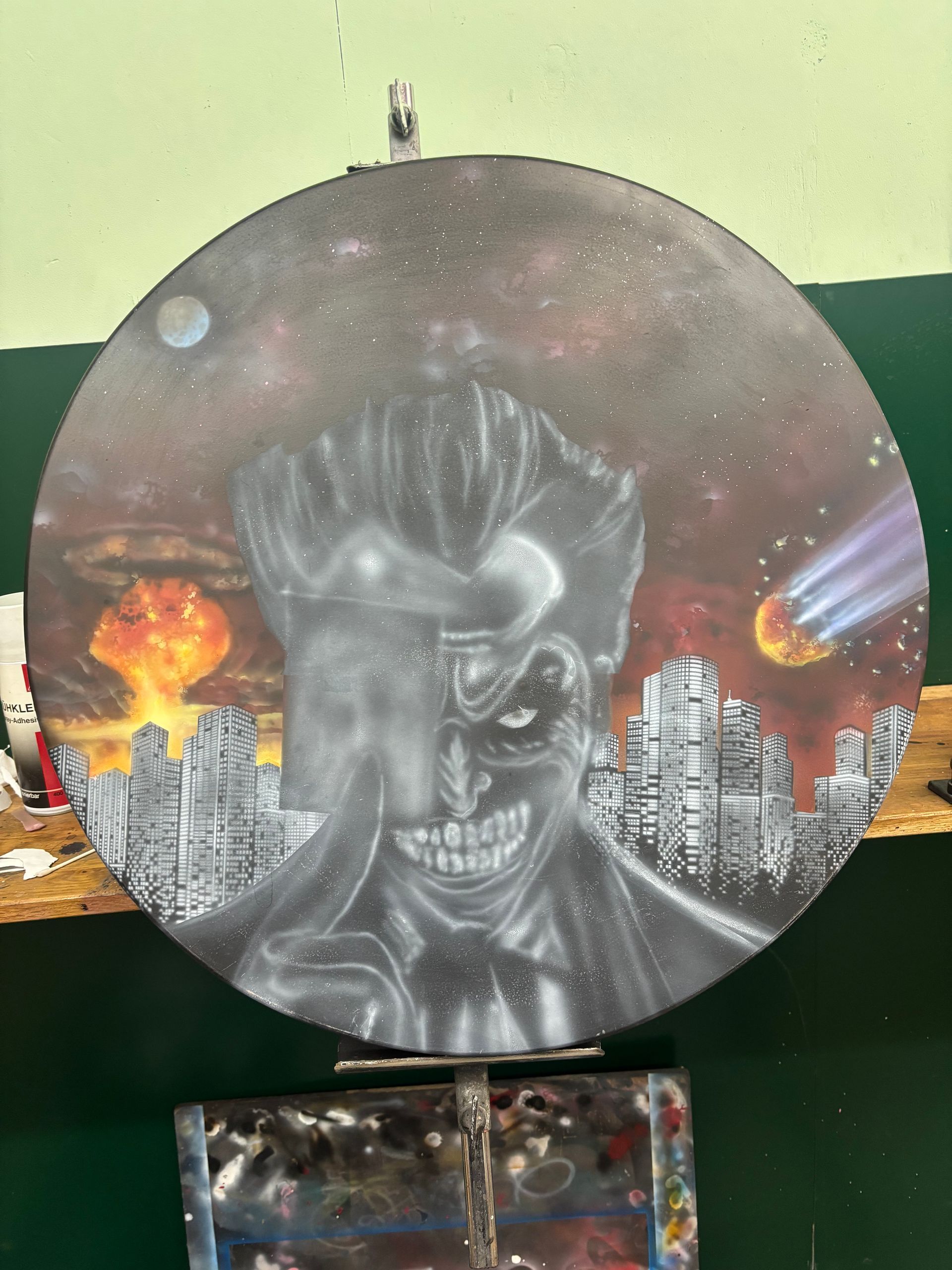 Rond houtskoolschilderij van een dreigend schedelgezicht met een vlammend stadslandschap en een donkere achtergrond.