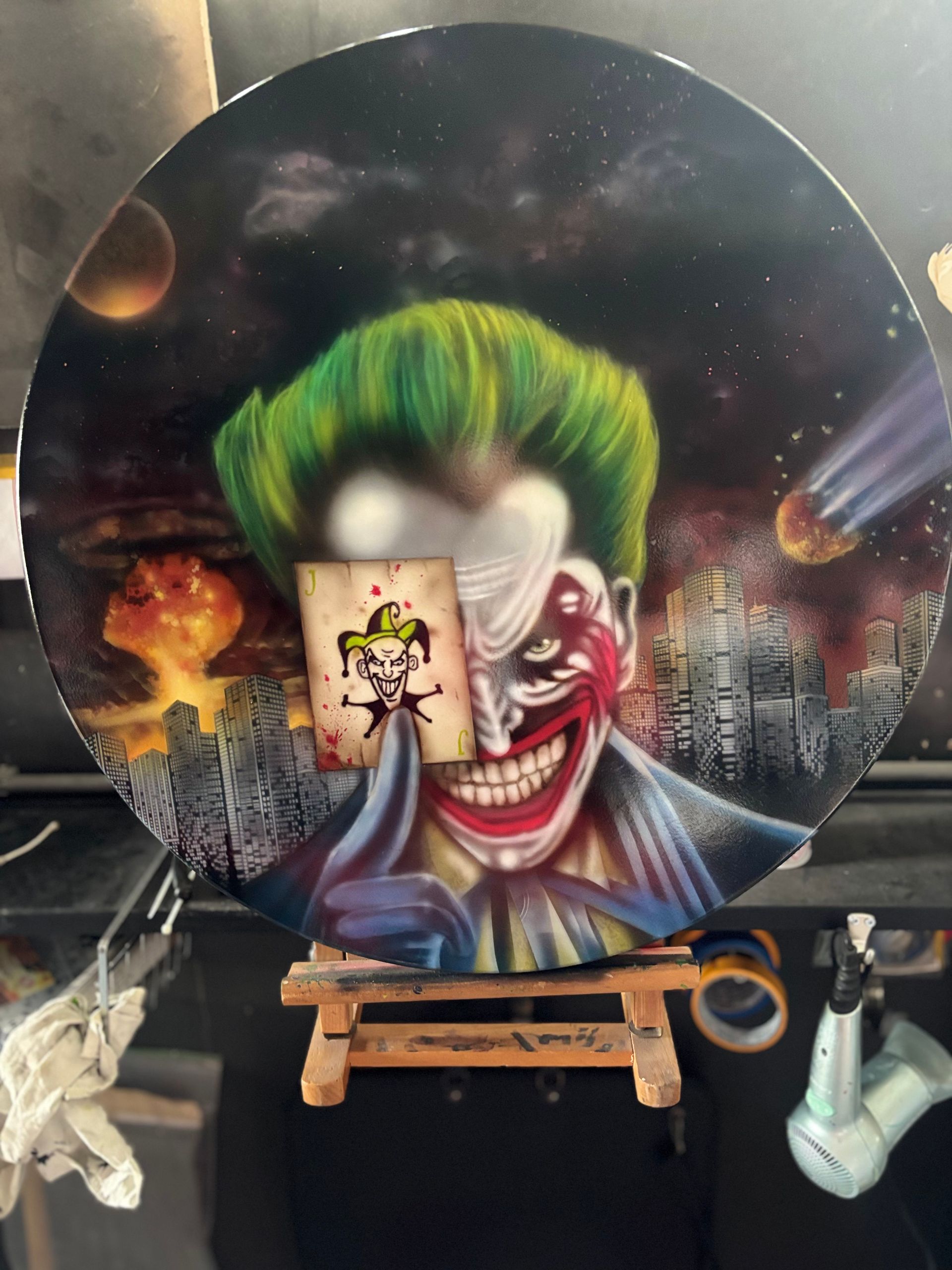Jokergezicht met groen haar op een rond doek, tentoongesteld op een houten schildersezel met een dramatische, rokerige achtergrond.