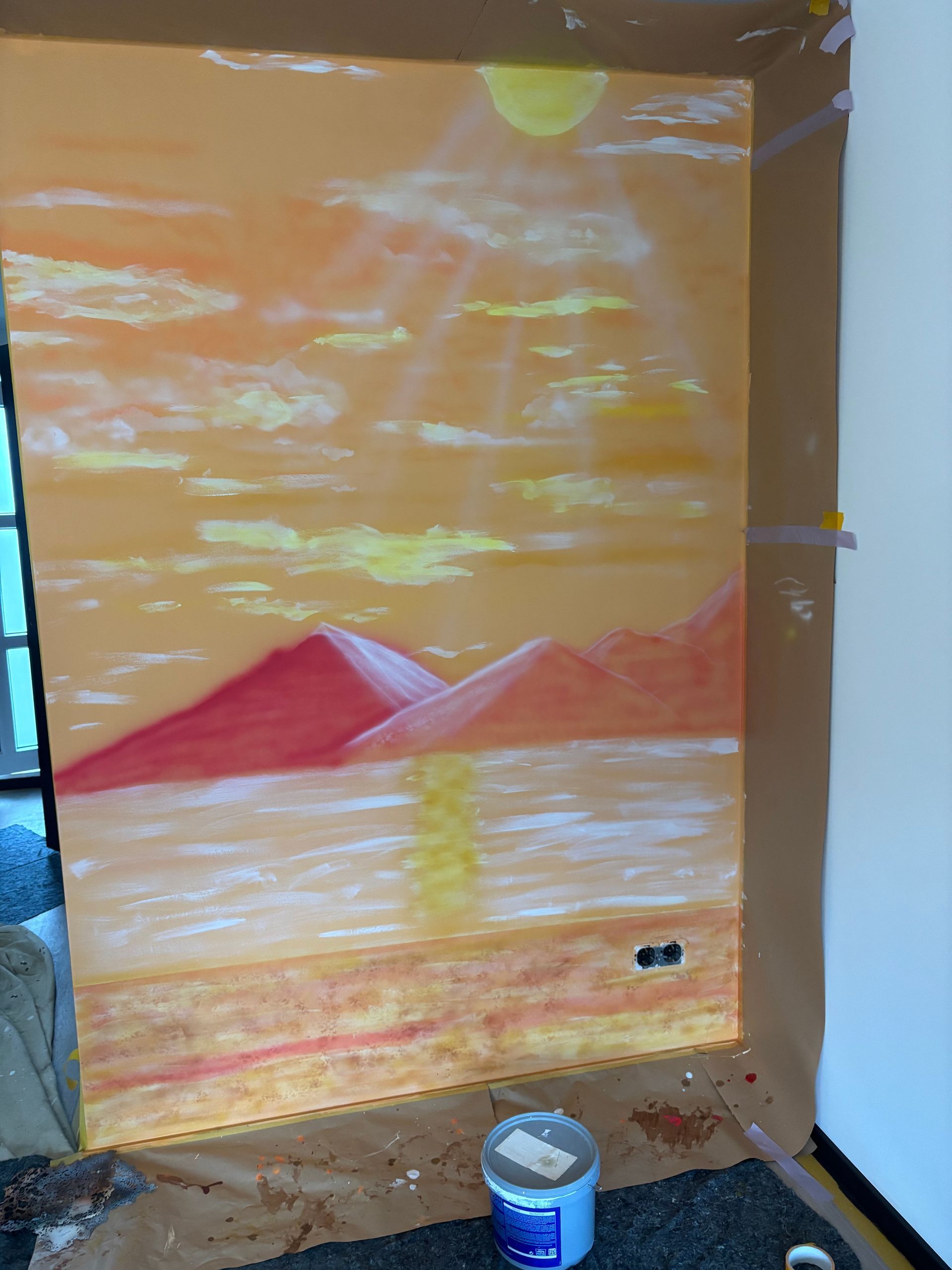 Een muurschildering van een oranje zonsondergang op een muur met rode bergen en reflecterend water, in een ruimte die momenteel wordt gerenoveerd.