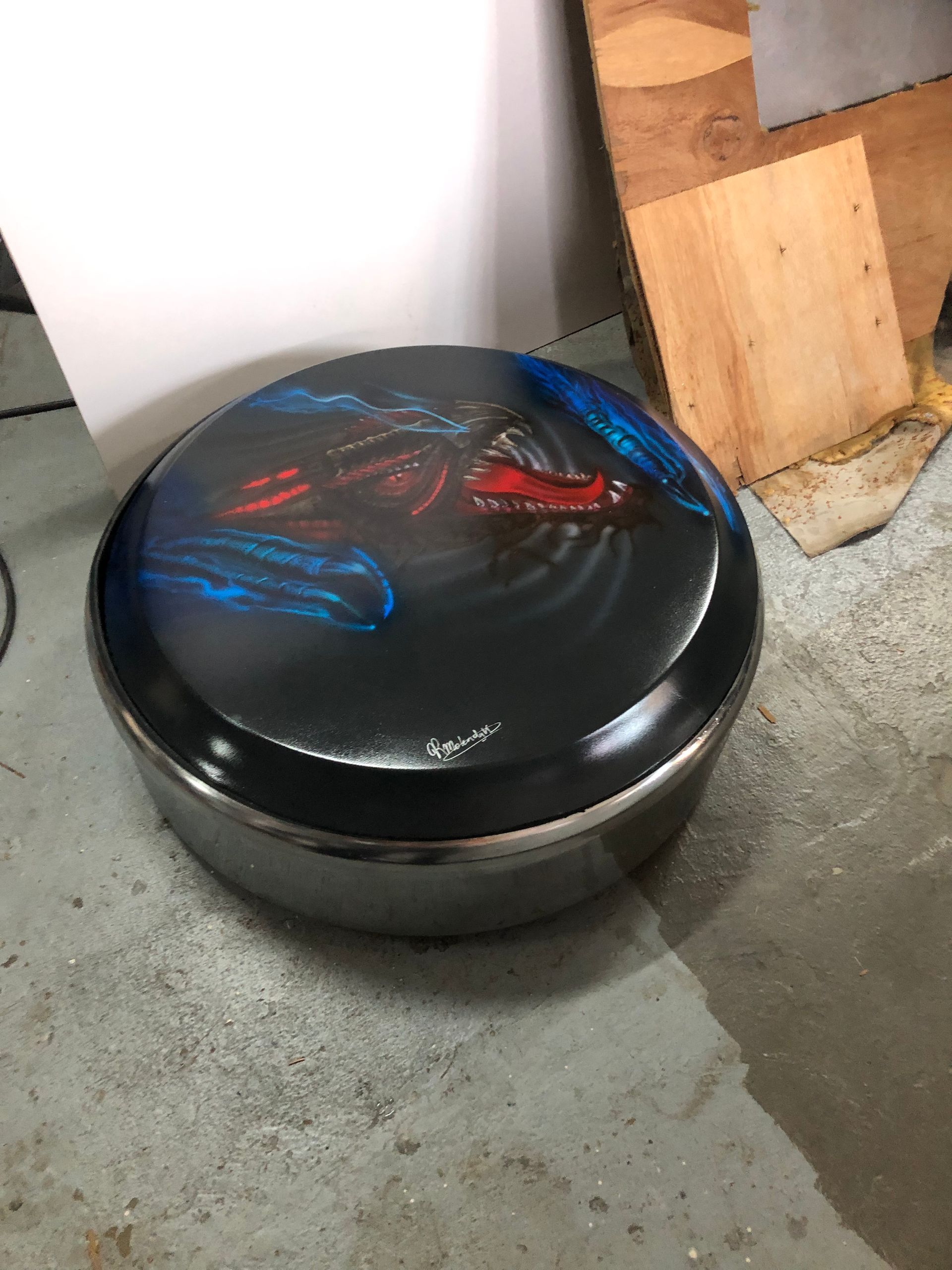 Zwarte ronde poef of kruk met een blauw en rood patroon op het blad, staand op een betonnen vloer naast houten planken.