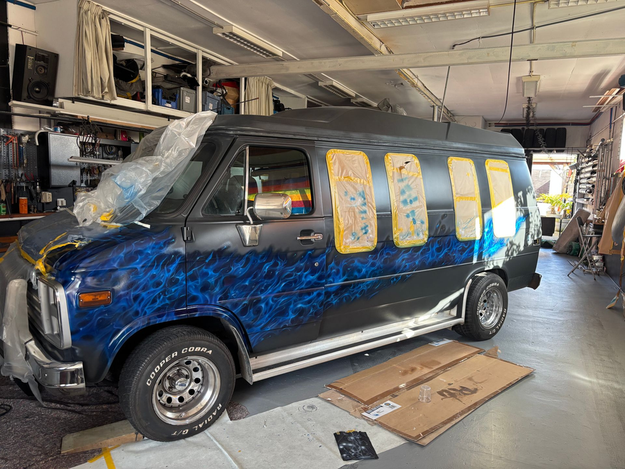 Blauwe bestelwagen met gele raamfolie geparkeerd in een garage, met gereedschap en apparatuur eromheen.