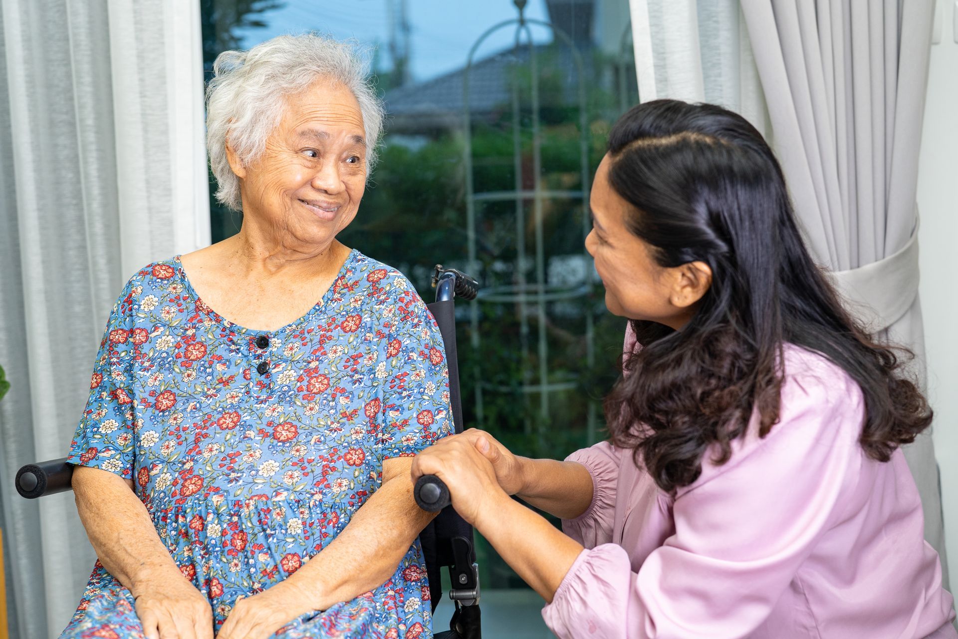 Woman in wheelchair smiles at woman holding her hand, near a window.