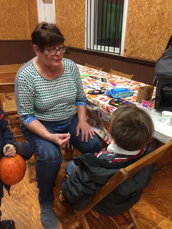 A woman is sitting in a chair talking to a child while face painting the child's face.