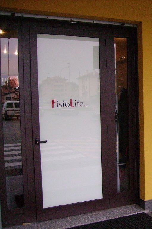 Ingresso di Fisiolife con logo sulla porta in vetro satinato. Telaio della porta marrone, parete gialla e attraversamento pedonale visibili.