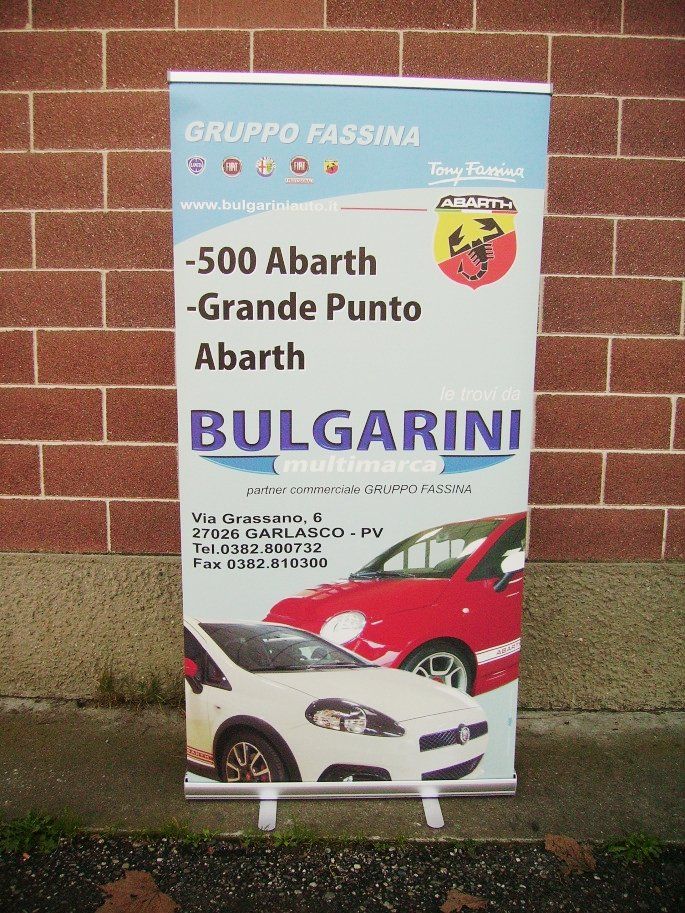 Banner roll-up per auto Abarth, esposto davanti a un muro di mattoni. Presenta immagini delle auto e informazioni aziendali.
