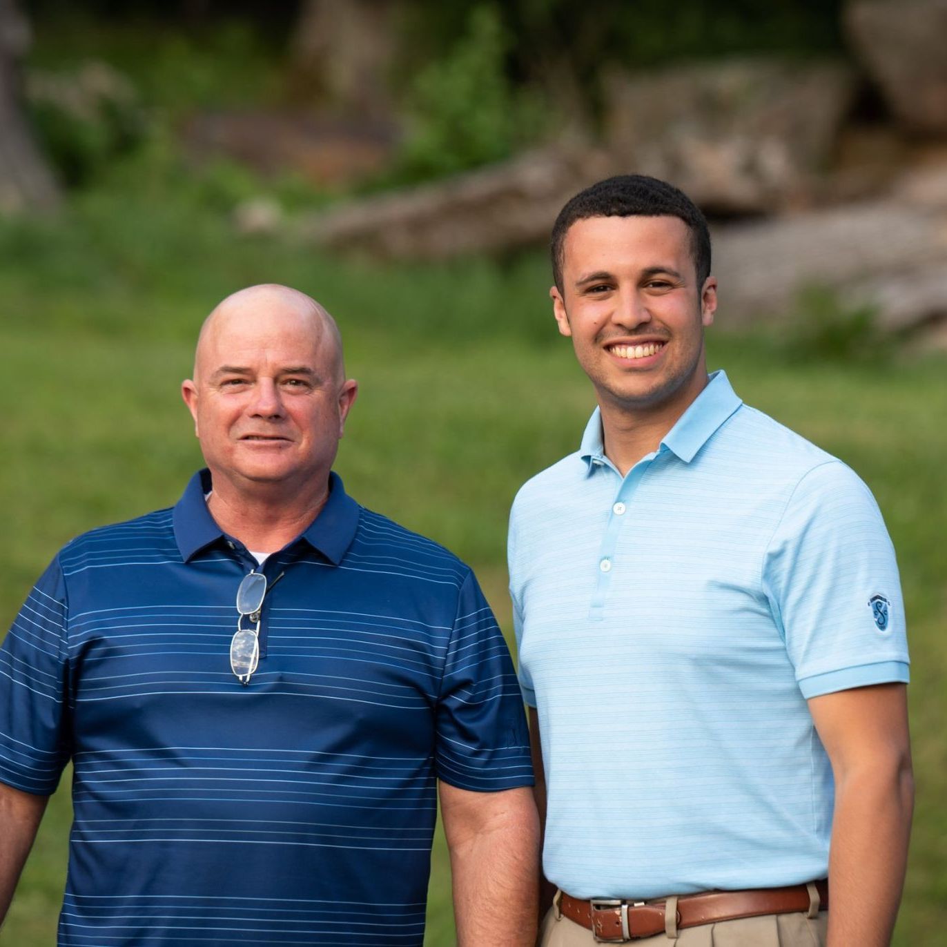 Two people standing side-by-side in a grassy outdoor setting. Both are wearing collared shirts and smiling at the camera.