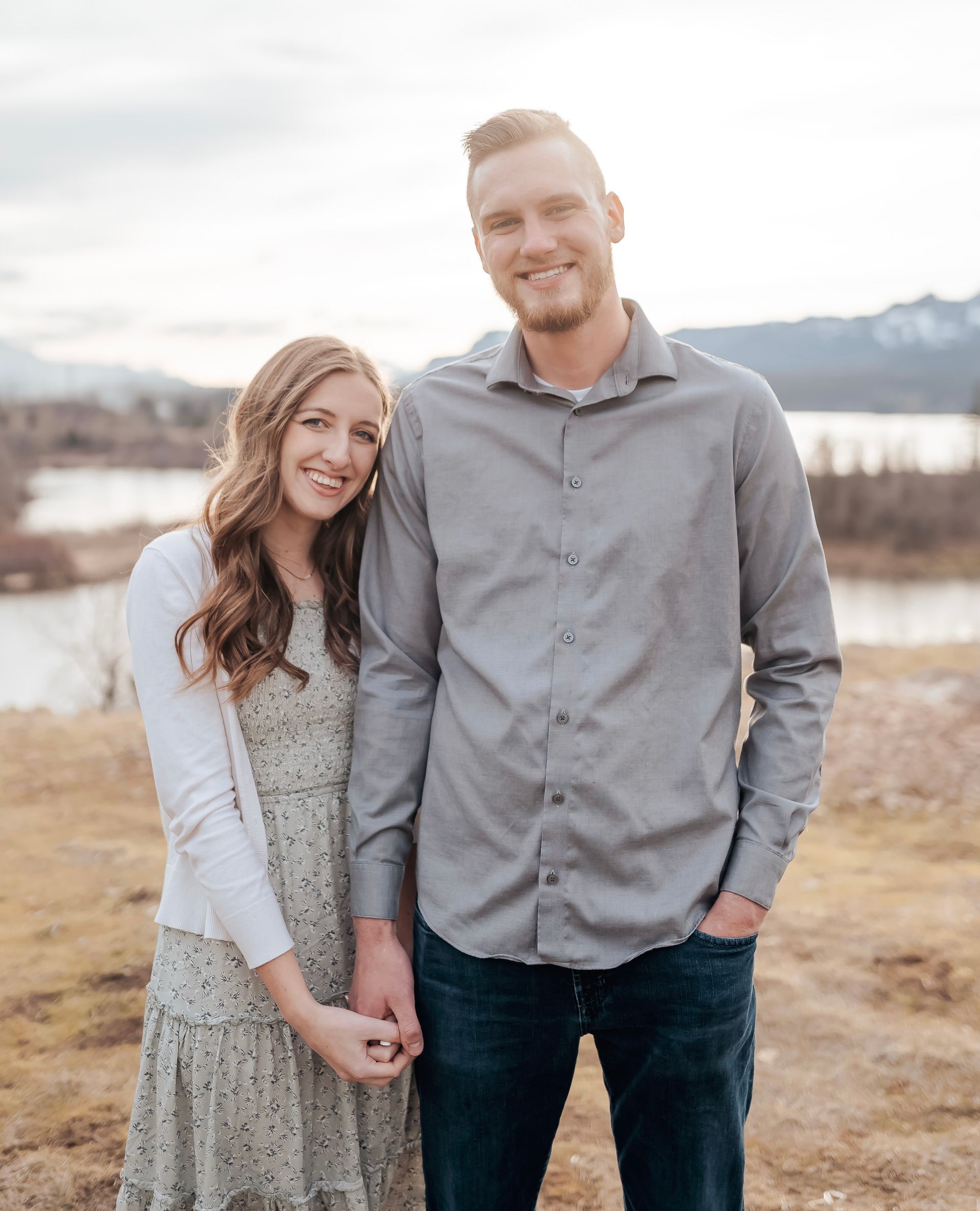 A couple stands close together, holding hands in a field near a river with mountains in the background.