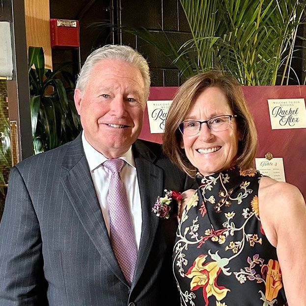 A person in a suit and tie stands smiling next to a person in a floral halter-neck dress in front of a decorative sign.