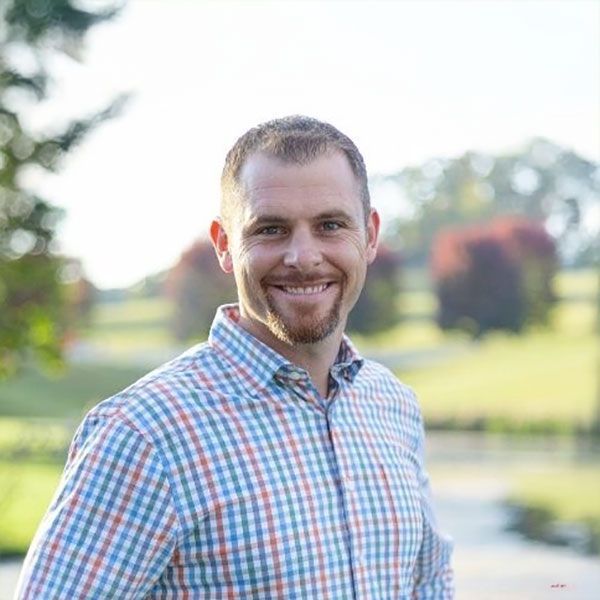 A smiling person with a short beard wears a blue, white, and orange plaid shirt, standing outdoors with trees in the back.