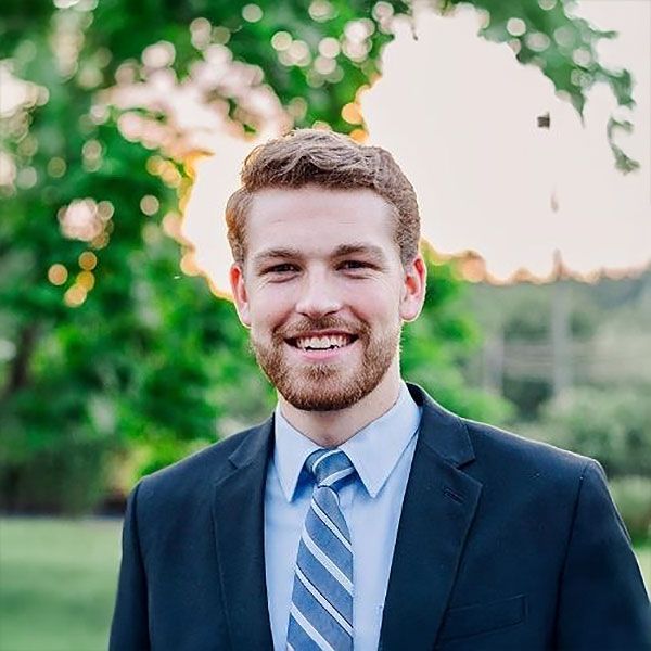 A person with a beard, wearing a navy suit, light blue dress shirt, and striped tie, smiling in front of a blurred garden.