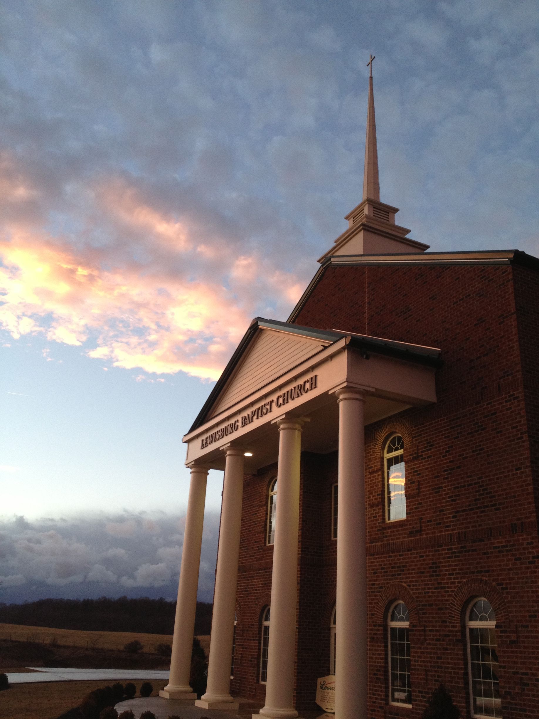 A brick church with white columns and a tall steeple against a cloudy, sunset sky.